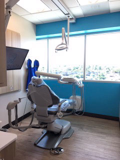 The image shows a dental office interior with modern equipment and furnishings, including a dental chair, a dental treatment table, and a large window allowing natural light into the room.