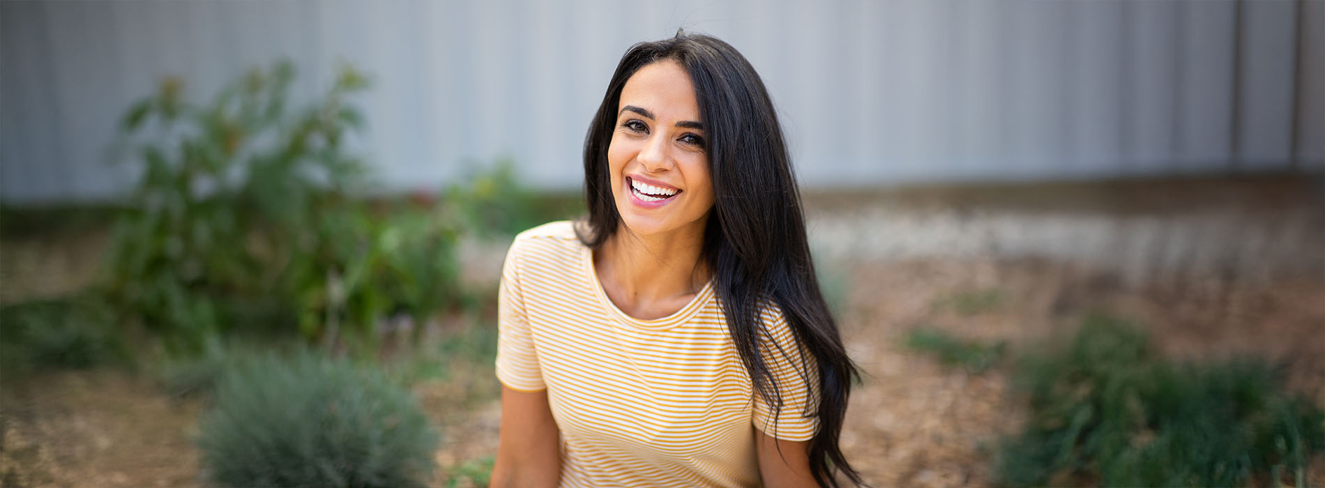 The image depicts a woman smiling at the camera, wearing a yellow top, standing outdoors with greenery in the background.