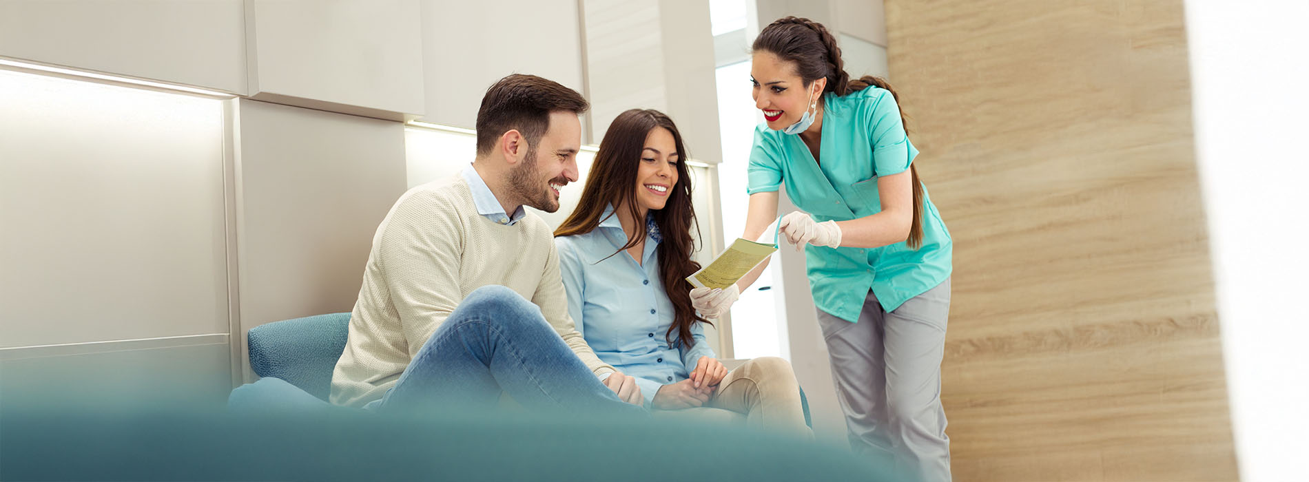 A group of people, including two standing individuals wearing blue scrubs, are gathered around a table with paperwork, smiling and engaged in conversation, with one person holding a clipboard and another sitting on a couch  they appear to be in an office setting.