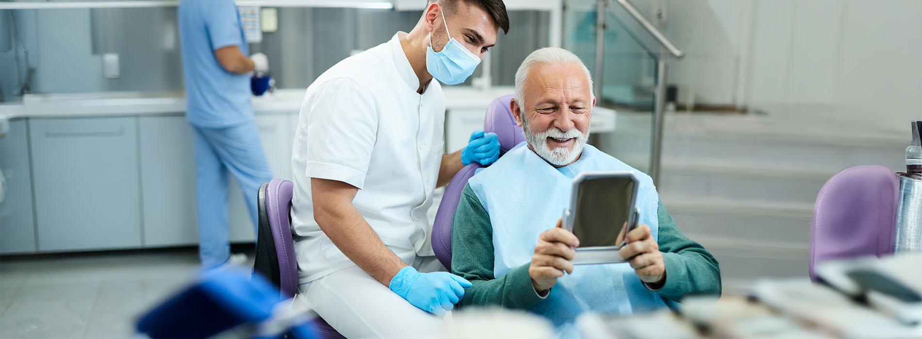 The image depicts two individuals in a dental setting, with one person seated at a desk holding a tablet, while another person stands over them wearing medical attire and a face mask.