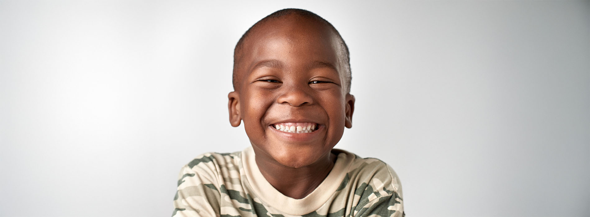 Young boy with a broad smile, wearing a camouflage shirt, against a plain background.