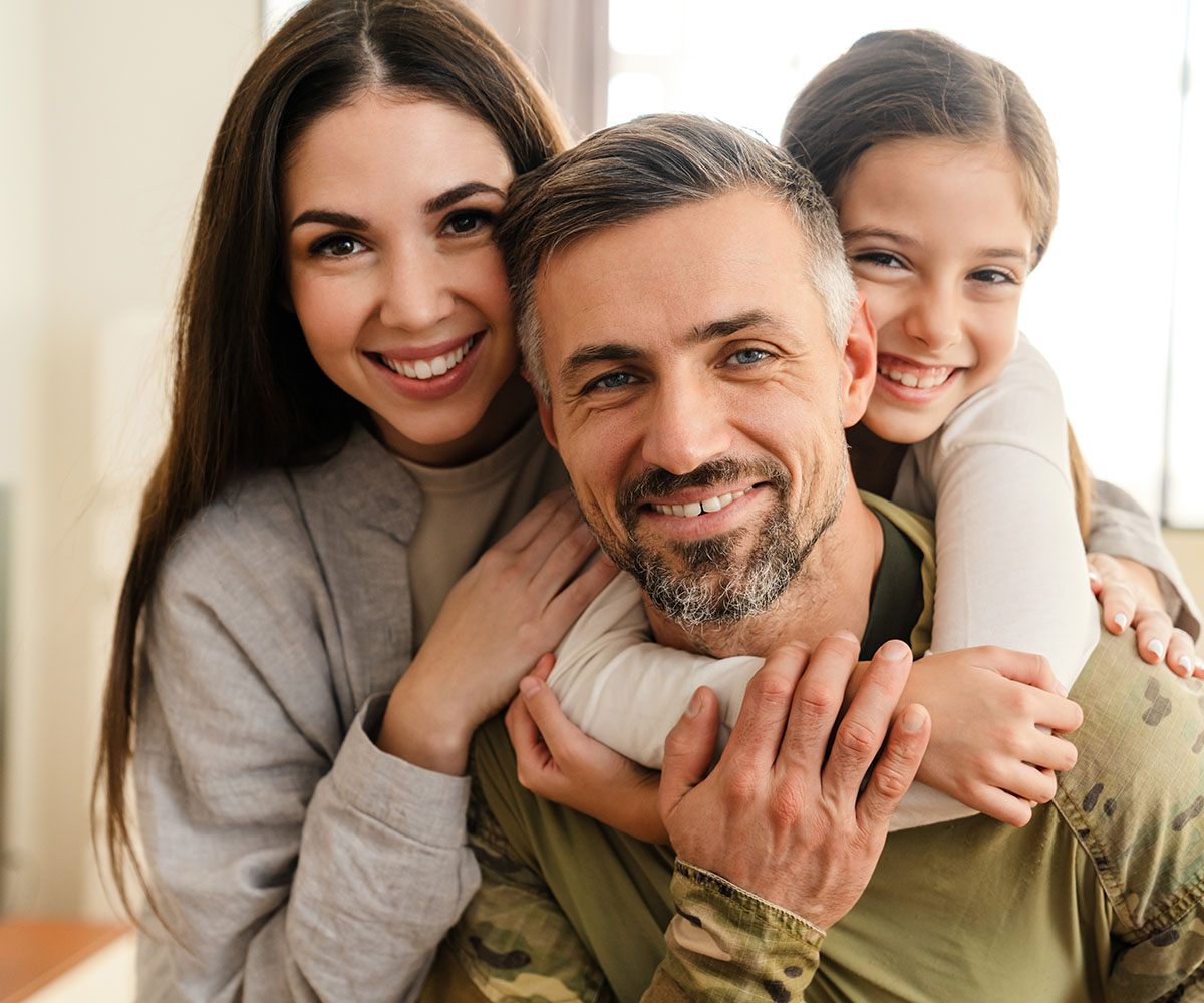The image shows a family with an adult man wearing military attire holding two children, with a woman embracing them all from behind.