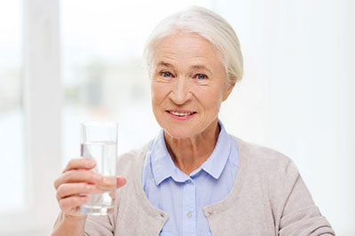 The image shows an elderly woman holding a glass of water with her right hand, smiling slightly, and looking directly at the camera.