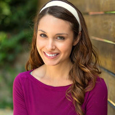 The image shows a woman smiling at the camera, wearing a purple top and a headband, with long hair, standing against a wooden fence in an outdoor setting.