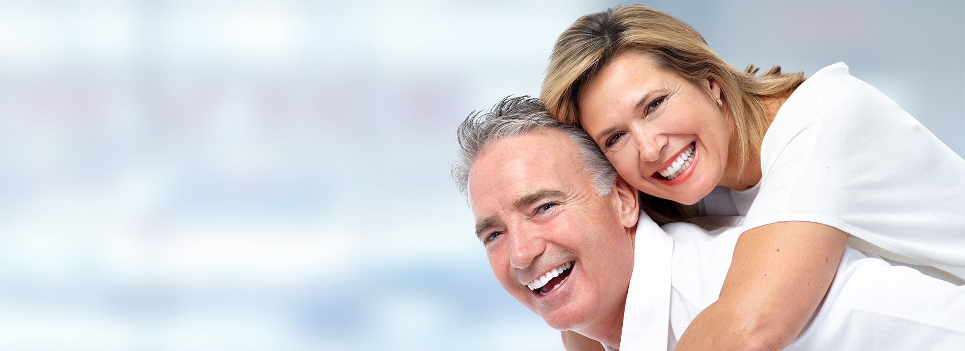 The image shows two adults, an older man and woman, smiling and embracing each other while posing for a photograph with a blurred background featuring blue and white hues.