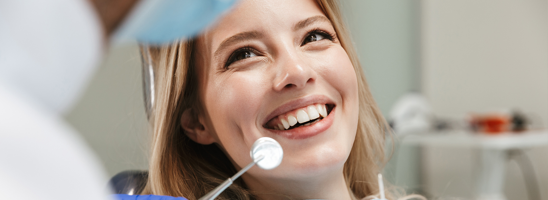 The image shows a woman with a smile, seated in front of a dental professional who appears to be holding a mirror during an examination.