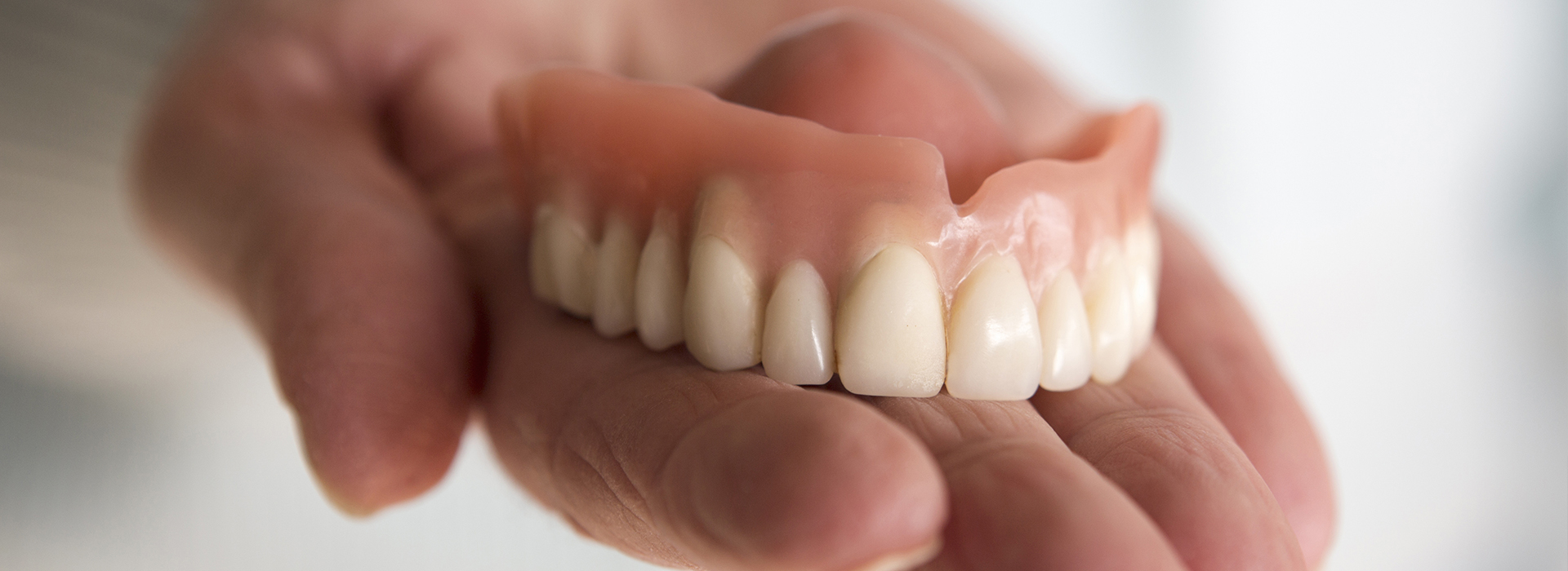 The image shows a hand holding a set of dentures with artificial teeth, against a blurred background.