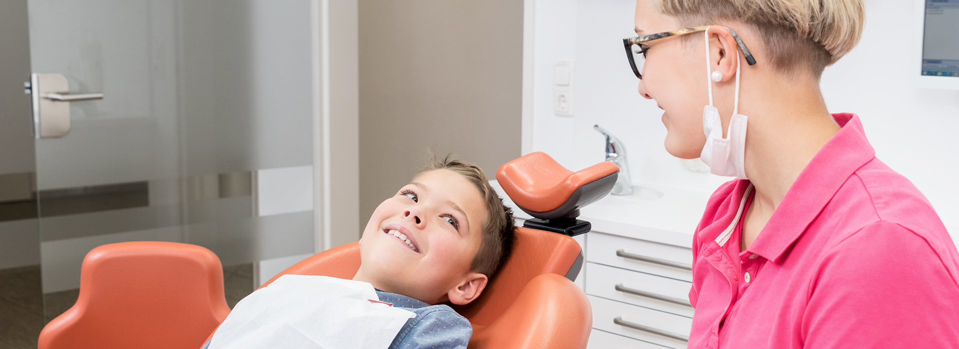 A woman sitting by a young boy who is laying on a dental chair with a smile on his face, receiving dental care from a professional seated behind him.