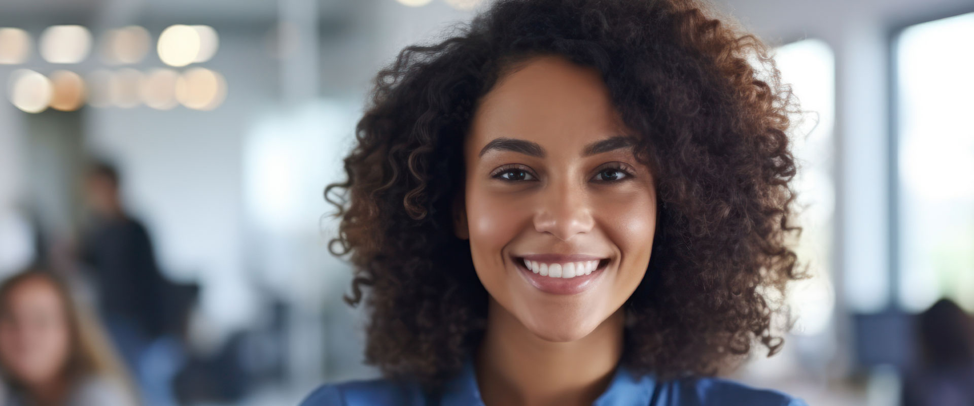 The image shows a smiling individual with curly hair, wearing a dark top, standing in an office environment with a blurred background featuring other people.