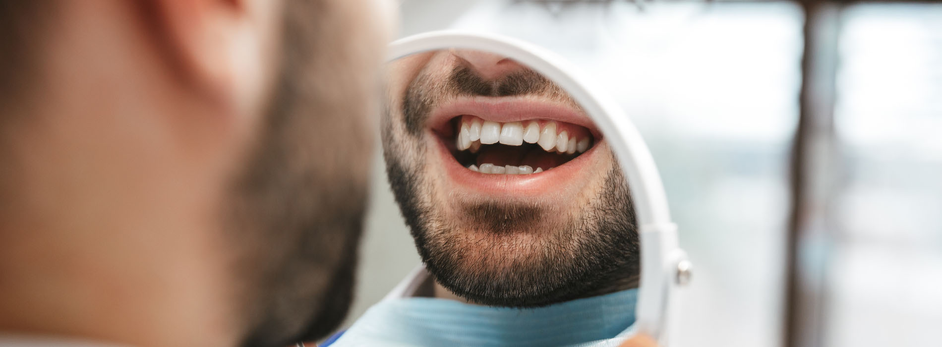 A man with a beard in a dental chair, smiling broadly while looking up at a dental mirror, with another man seated beside him, possibly a dentist, wearing a face mask and stethoscope.