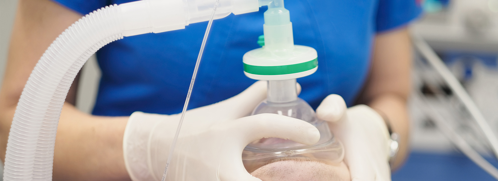 The image shows a person wearing gloves and a blue shirt, standing at a counter with equipment that includes a transparent plastic tube connected to a green device, which appears to be a medical or scientific instrument, possibly related to a laboratory setting.