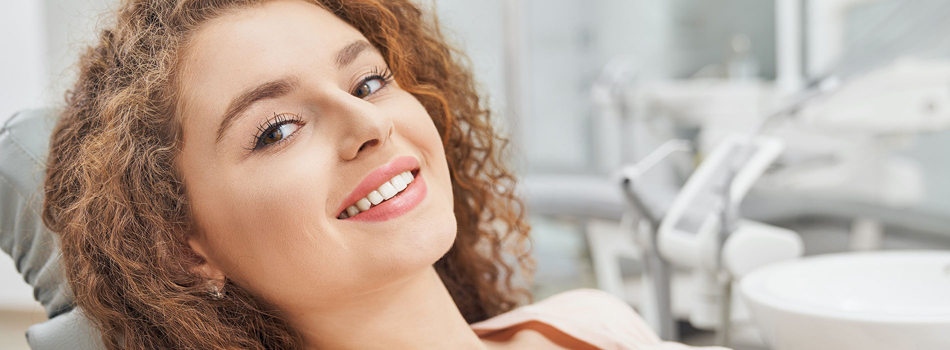 The image shows a young woman with curly hair smiling at the camera while sitting in a dental chair.