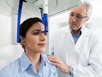 The image shows a person sitting in a dental chair with a blue dental device attached to their head, being assisted by a dental professional who appears to be a dentist.