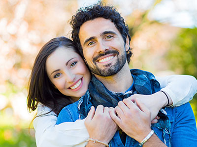 A man and woman are embracing each other with a smile, set against a blurred background.