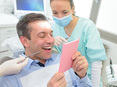 The image depicts a man sitting in a dental chair with a smiling expression, holding a pink card with text on it, while a woman wearing a surgical mask stands behind him, looking at the card with a slight smile.