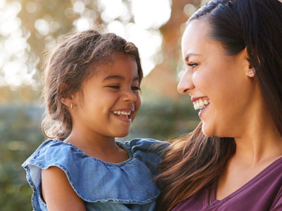 The image depicts a woman smiling at her child while holding them, both outdoors with trees in the background.