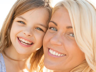 The image depicts a woman and a young girl, both smiling at the camera, with the woman appearing to be an adult and the child being a little girl. They are outdoors under bright sunlight, likely on a sunny day, given the shadows and lighting conditions. The woman has blonde hair and is wearing a light-colored top, while the child has brown hair and is dressed in a sleeveless top with darker tones.