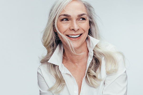 The image shows a woman with short blonde hair smiling at the camera. She has gray hair on top and is wearing a white shirt.