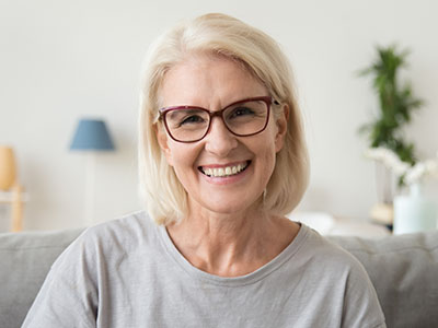 A smiling woman with short blonde hair wearing glasses and a light-colored top, seated indoors with a homey background.