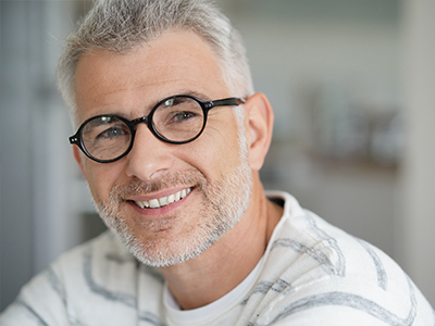 The image shows a middle-aged man with short gray hair, wearing glasses, smiling at the camera, with a beard and mustache, dressed in a white shirt with a patterned collar.