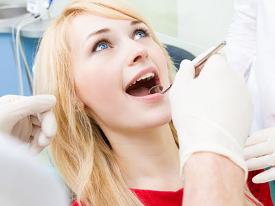 A woman sitting in a dental chair with her mouth open, receiving dental care from a dentist who is examining her teeth while wearing gloves and a face mask.