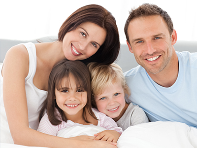 A family of four posing on a bed with smiles, including two adults and two children.