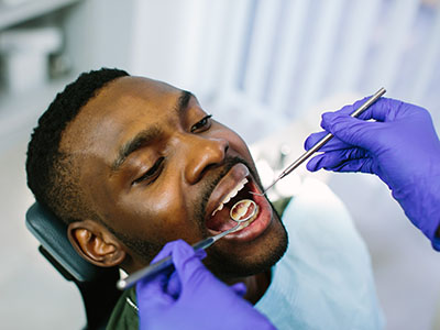 The image shows a person receiving dental care, with a dentist performing work on their teeth using dental tools while wearing protective gloves.