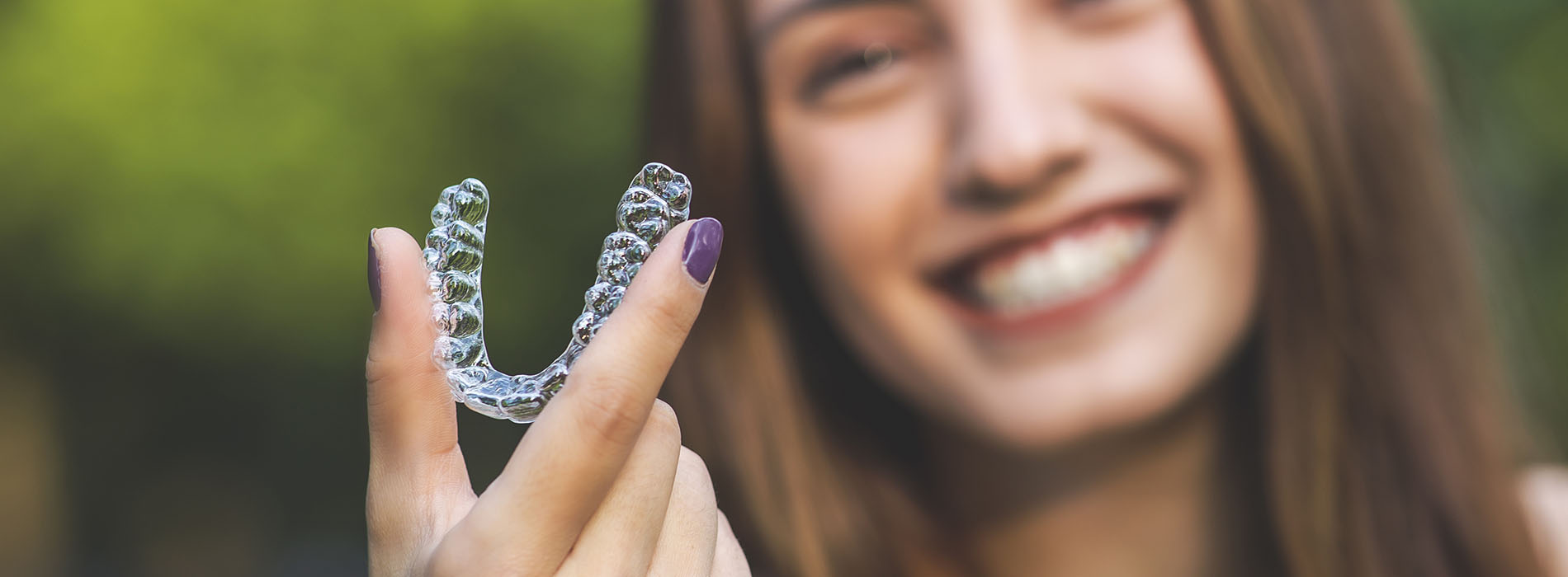 A smiling woman holding up a clear plastic dental retainer with her left hand.